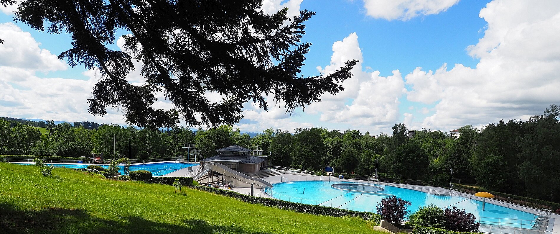 Ein Schwimmbad unter freiem Himmel mit mehreren Wasserbecken. Grünflächen und Bäume umgeben den Poolbereich, während ein klarer blauer Himmel mit einigen Wolken zu sehen ist. Die Umgebung ist ruhig und einladend für Badegäste.