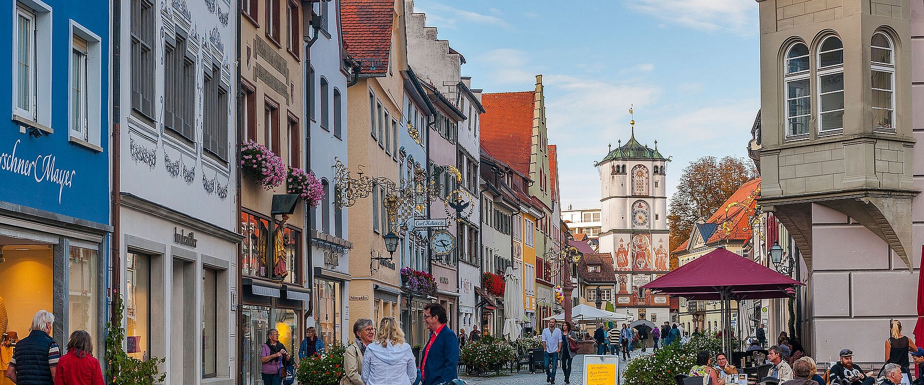 Eine belebte Straße in einer Stadt mit historischen Gebäuden und bunten Fassaden. Menschen gehen entlang der Promenade, es gibt Tische unter Sonnenschirmen. Im Hintergrund ist ein Uhrturm zu sehen, umgeben von Bäumen und einem klaren Himmel.