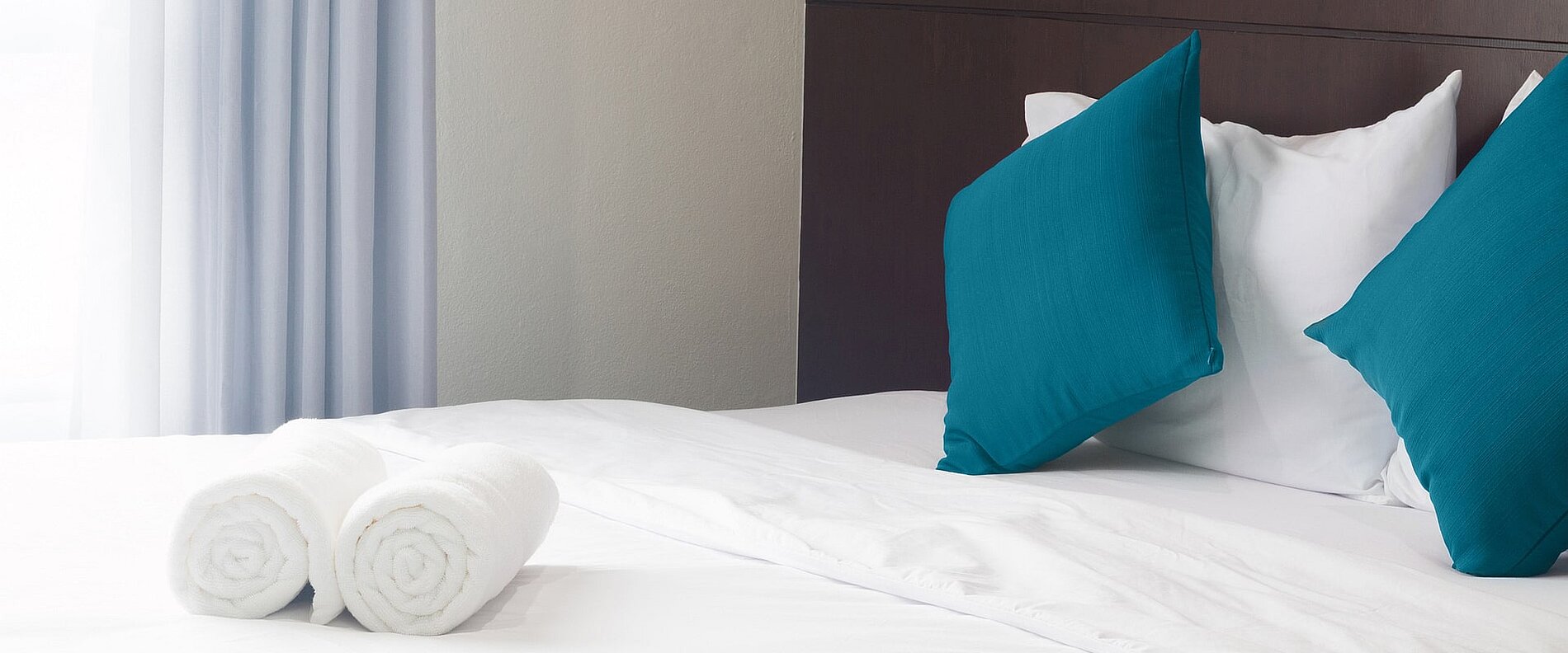 Ein frisch bezogenes Bett mit weißer Bettwäsche, geschmückt mit zwei türkisfarbenen Kissen. Auf dem Bett liegen zwei gerollte, weiße Handtücher. Licht strömt durch ein Fenster mit hellen Vorhängen.