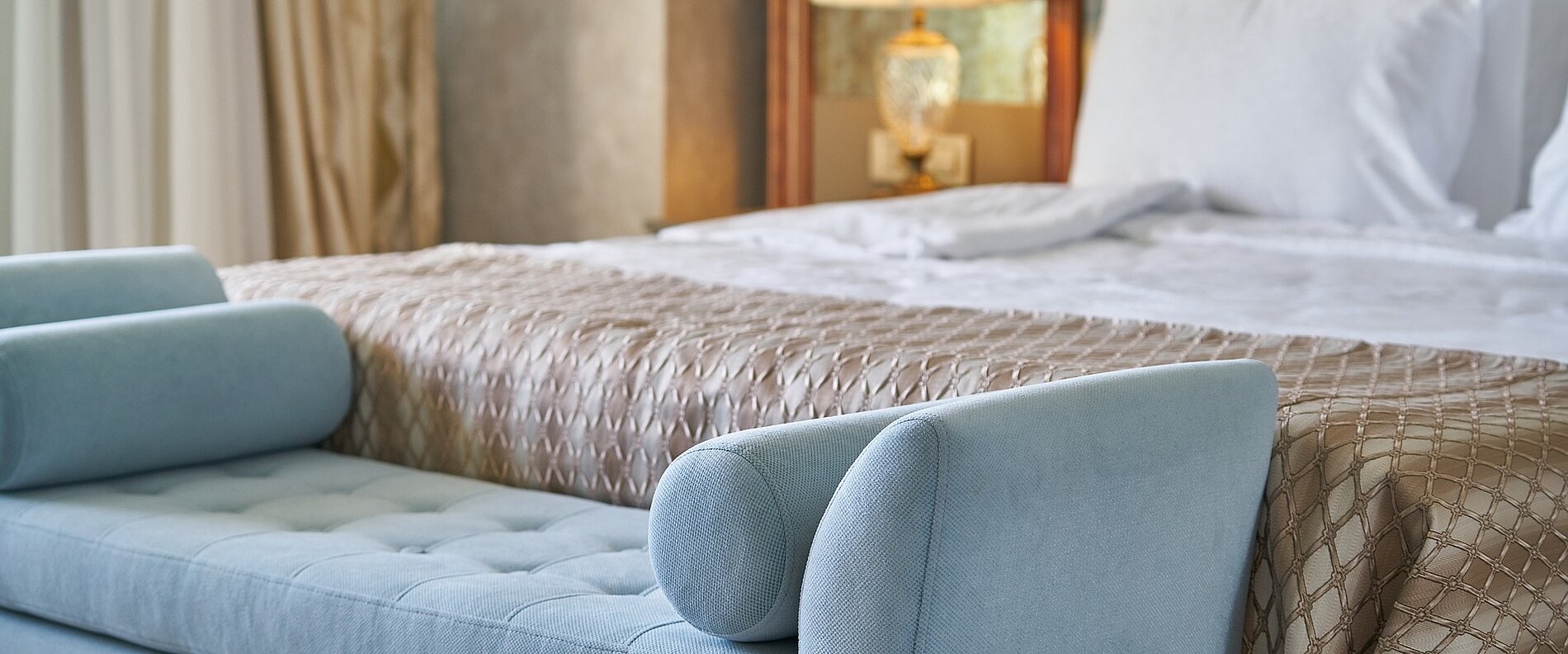 Ein elegantes Schlafzimmer mit einem großen Bett, das mit einer goldfarbenen Decke dekoriert ist. Am Fußende des Bettes steht eine blaue Bank mit zwei runden Kissen. Im Hintergrund sind Vorhänge und eine Lampe sichtbar.