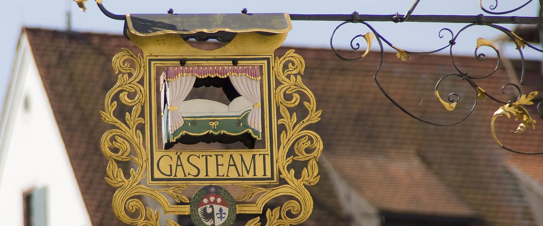 Ein bunter Wirtshausschild mit der Aufschrift "Gästeam" und einem stilisierten Bett, umgeben von dekorativen Verzierungen. Der Hintergrund zeigt ein Gebäude mit einem Dach und Fenstern.