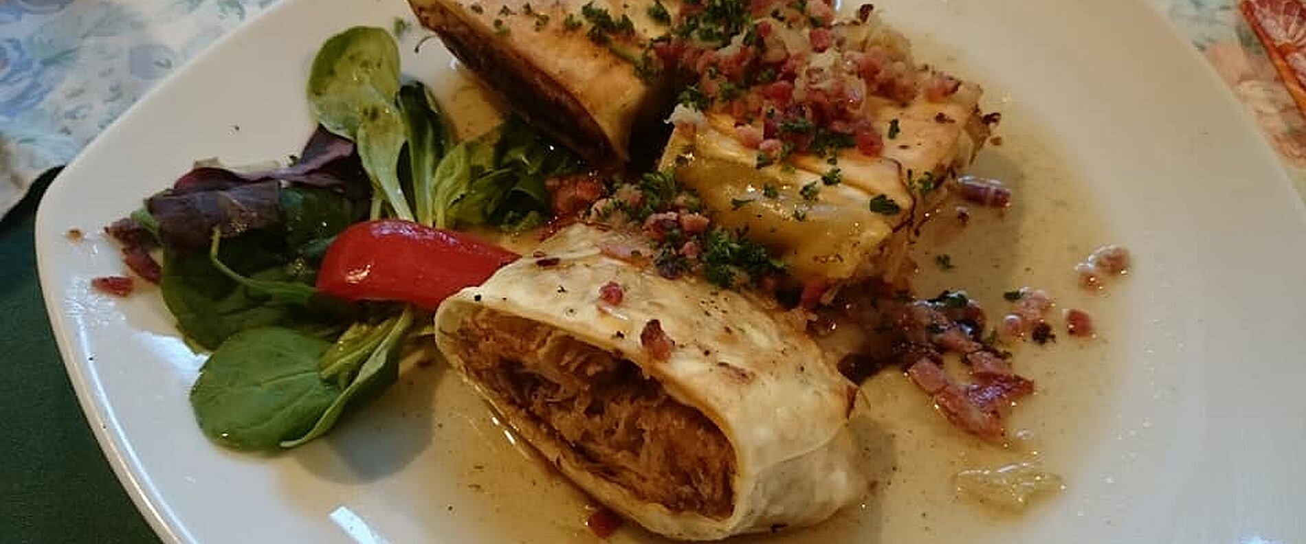 Ein Teller mit gefülltem Kohl, umgeben von einem kleinen Salat aus grünen Blättern und Paprika. Das Gericht ist mit Kräutern und Speckstückchen garniert und in einer Sauce angerichtet.