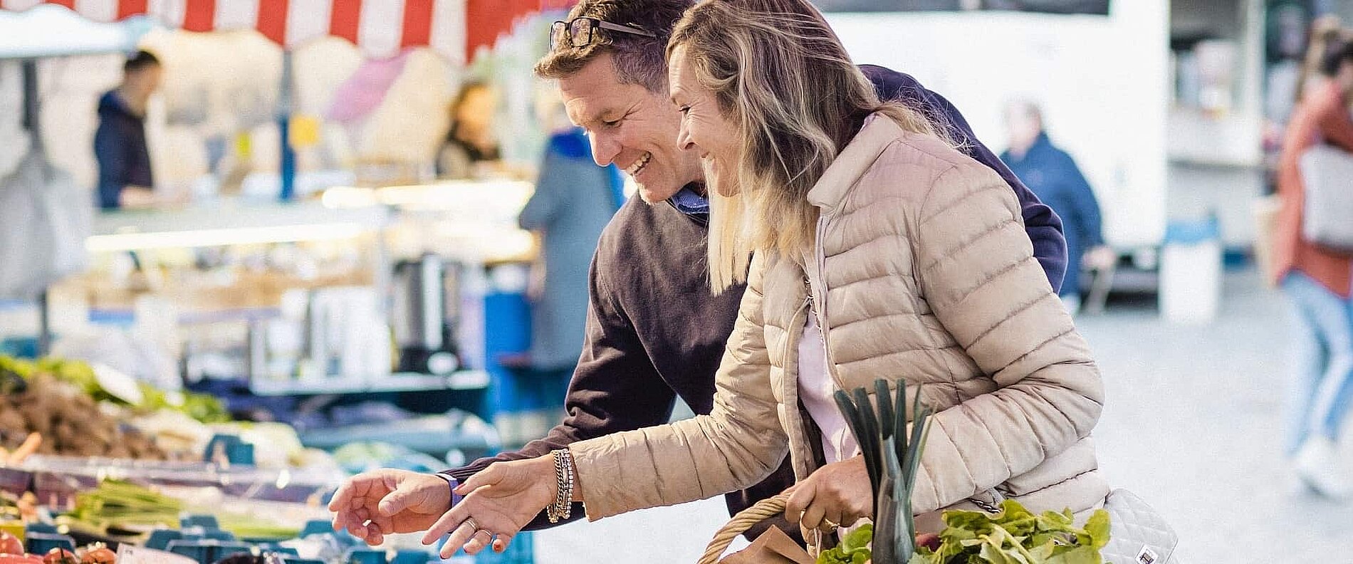 Ein Paar steht auf einem Markt und betrachtet frisches Gemüse. Die Frau hält einige Gemüsearten in den Händen, während der Mann lächelt und sie anspricht. Im Hintergrund sind weitere Marktstände und Menschen zu sehen.