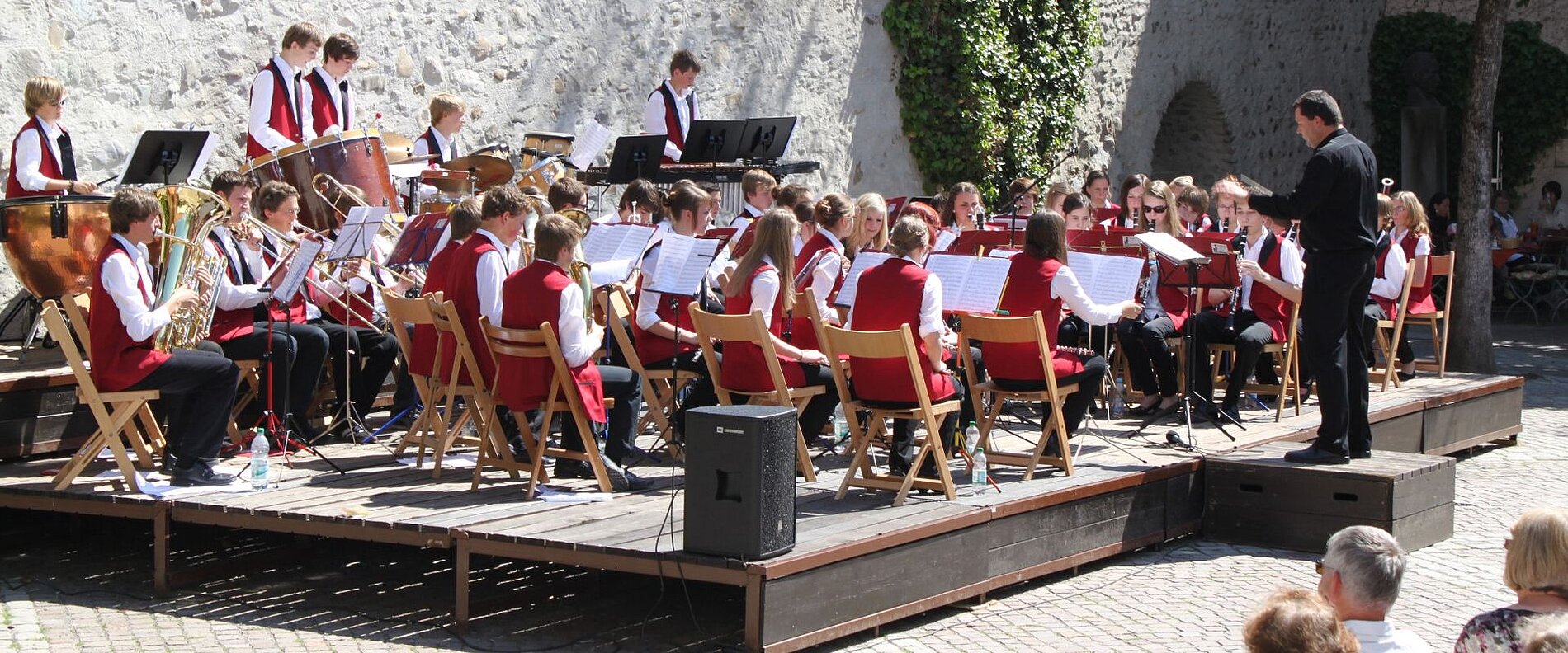 Eine Blaskapelle spielt auf einer Freiluftbühne. Die Musiker tragen rot-weiße Kleidung und sind in mehreren Reihen angeordnet. Ein Dirigent leitet die Aufführung. Im Hintergrund sind Menschen zu sehen, die das Konzert beobachten.