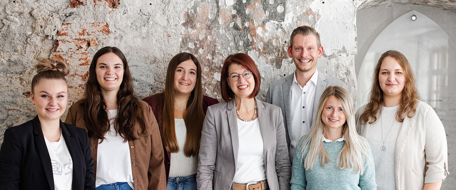 Eine Gruppe von acht Personen steht vor einer rauen Steinwand. Sie lächeln und tragen verschiedene professionelle outfits. Die Personen sind gemischt zwischen Männern und Frauen, wobei einige lange Haare haben und andere kürzere Frisuren.