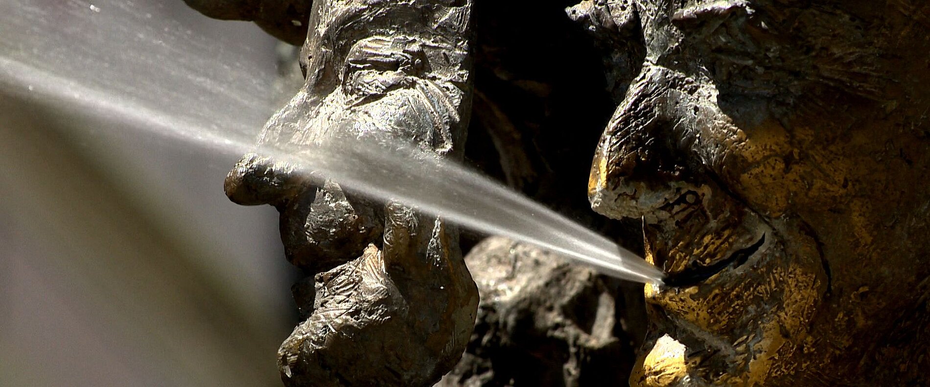 Die Nahaufnahme einer Skulptur zeigt zwei Gesichter, die sich aneinanderneigen. Aus einer Öffnung zwischen ihnen spritzt Wasser, was einen dynamischen und interaktiven Effekt erzeugt. Die Gesichter sind detailreich und stilisiert, mit einer Betonung auf den Ausdruck.